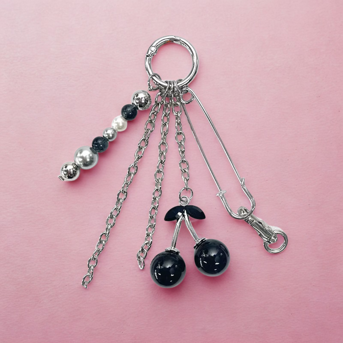 Llavero con cerezas negras brillantes y gancho metálico, sobre fondo rosa claro. Accesorio decorativo con estilo pop y original.