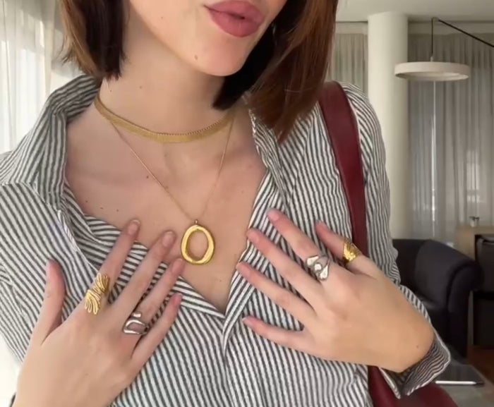 Collar dorado con dije irregular orgánico grande sobre fondo claro y anillos CARLOTA sobre modelo