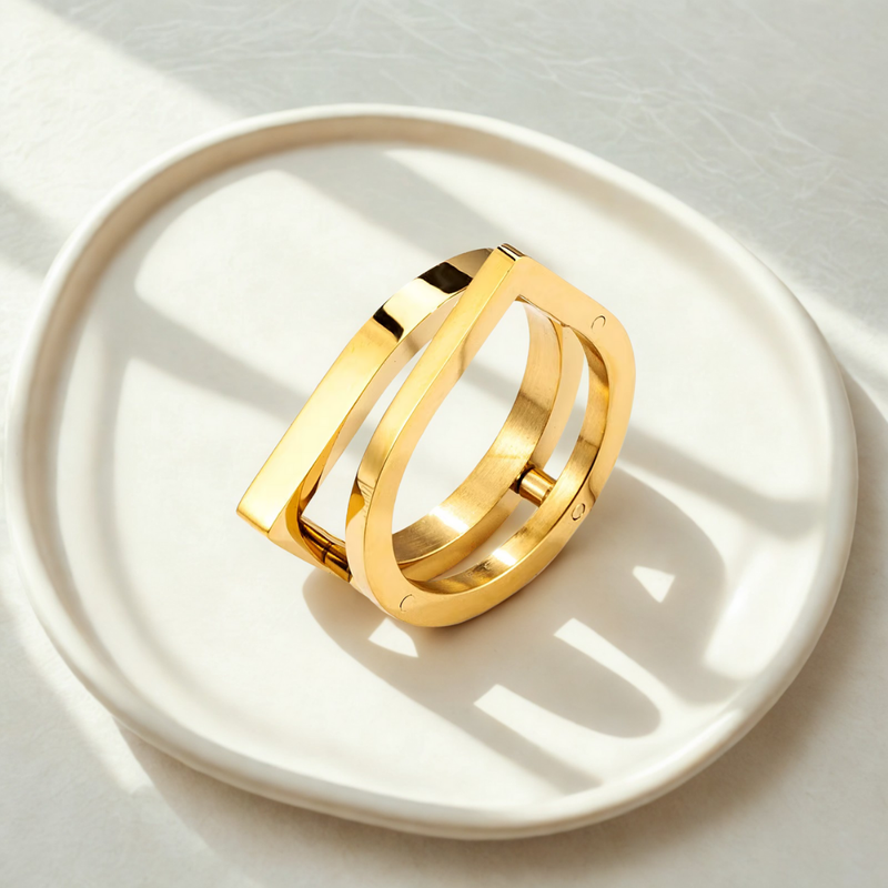 Anillo dorado de acero quirúrgico con forma arquitectónica moderna sobre bandeja blanca.