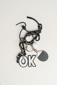 Llavero paddle en metal plateado, cordón negro y simbolo de OK. Ideal para llaves o como accesorio de estilo.