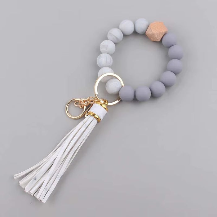 Llavero tipo pulsera con cuentas en tonos de gris, detalle de cuenta de madera y borla blanca de flecos de cuero sintético.