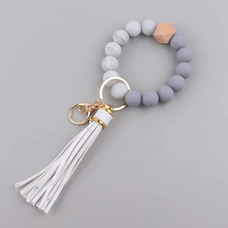 Llavero tipo pulsera con cuentas en tonos de gris, detalle de cuenta de madera y borla blanca de flecos de cuero sintético.