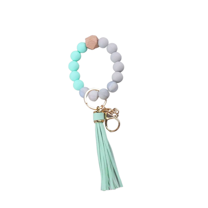 Llavero tipo pulsera con cuentas en blanco y verde agua, detalle de cuenta de madera y borla de flecos de cuero sintético de verde claro.