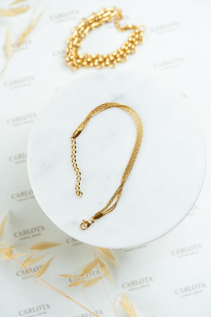 Pulsera dorada de varias cadenas finas en acero quirúrgico y pulsera gruesa de fondo desenfocada de CARLOTA